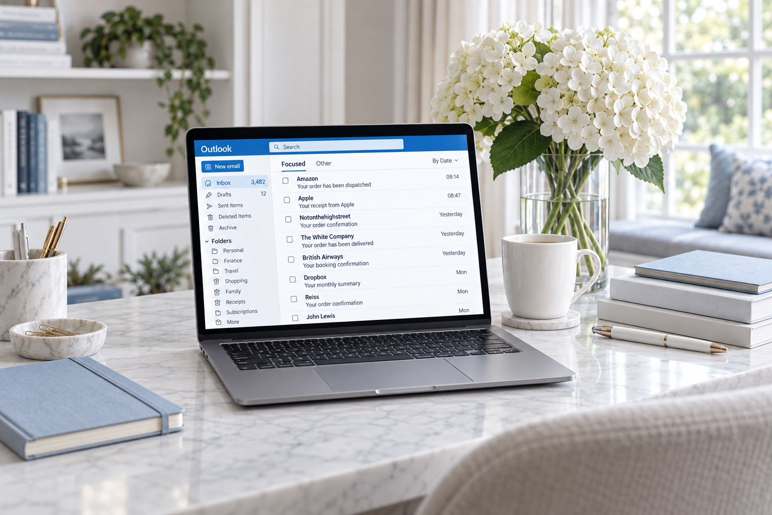 A landscape photograph of an Outlook inbox on a laptop beside white hydrangeas, a mug and pale blue notebooks.