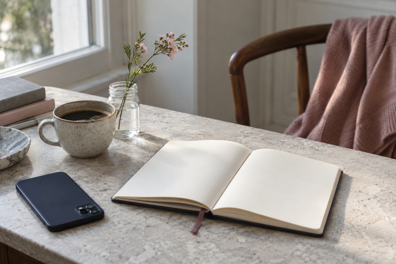 An open blank notebook, face-down phone, coffee and small pink flowers on a pale stone table.